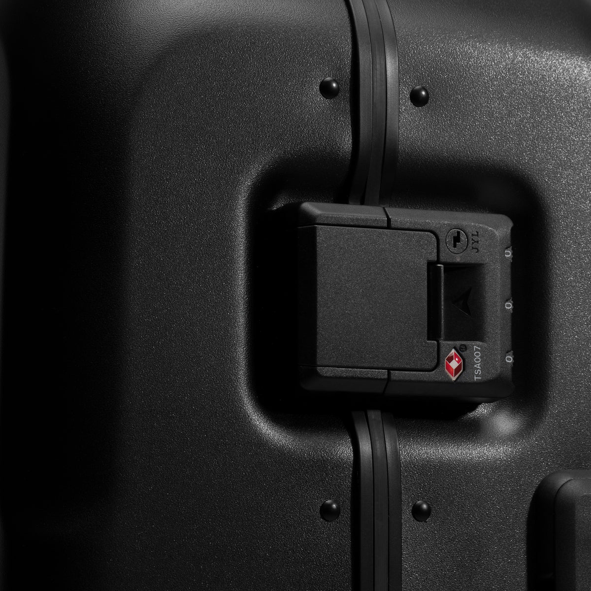Close-up of a black hard-shell suitcase showing the luggage lock mechanism and a small red diamond-shaped logo on the lock cover. [All variants]