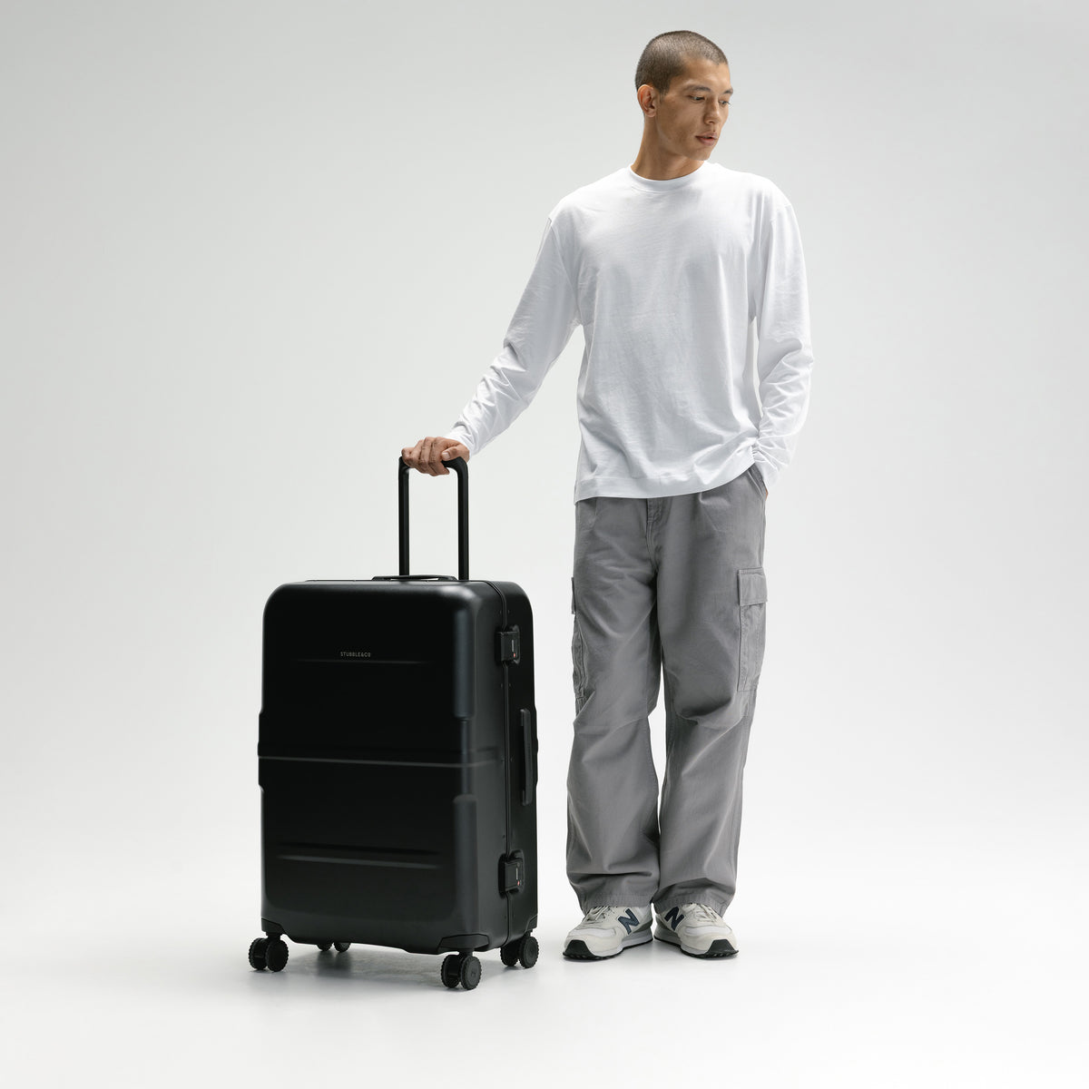 A man in a white long-sleeve shirt and gray cargo pants stands next to a black hard-shell rolling suitcase with its telescoping handle extended, against a light gray studio background. [All variants]