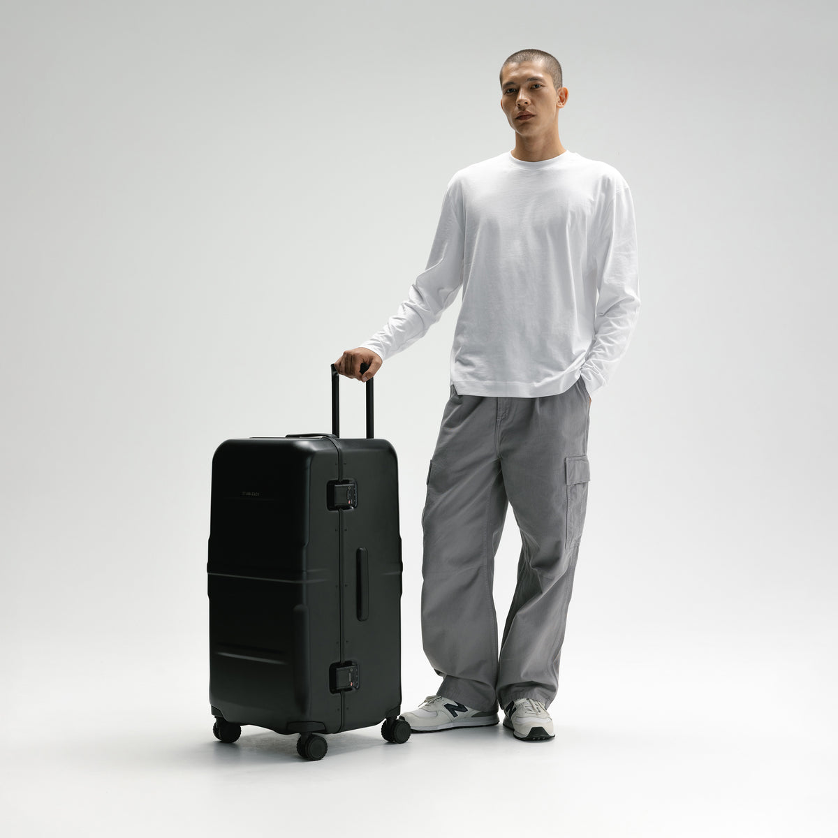 A man in a white long-sleeve shirt and gray cargo pants stands next to a black hard-shell rolling suitcase, holding its telescoping handle in a studio setting with a light gray background.  [All variants]