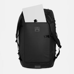 All Black duffel backpack shown from the front on a light gray background, with a silver laptop partially inserted into the top opening. A small white geometric logo is visible on the front pocket.