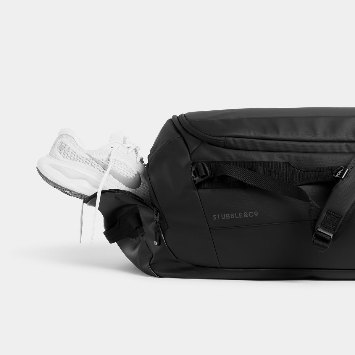 All Black duffel bag with a white sneaker partially tucked into the side pocket, photographed on a clean light gray studio background.