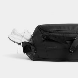 All Black duffel bag with a white sneaker partially tucked into the side pocket, photographed on a clean light gray studio background.