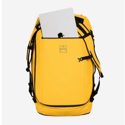 Yellow duffel backpack with black straps and trim, shown on a light gray background with a silver laptop partially tucked into the top pocket. A small geometric logo is visible on the front.