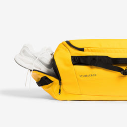 Yellow duffel bag with black straps and a white sneaker partially tucked into its side zipper, shown against a clean white studio background.