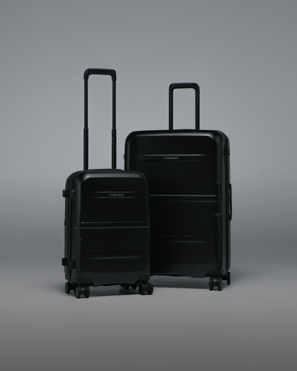 Two hard-shell spinner suitcases in matte black, one small and one large, standing upright on a smooth gray studio background.