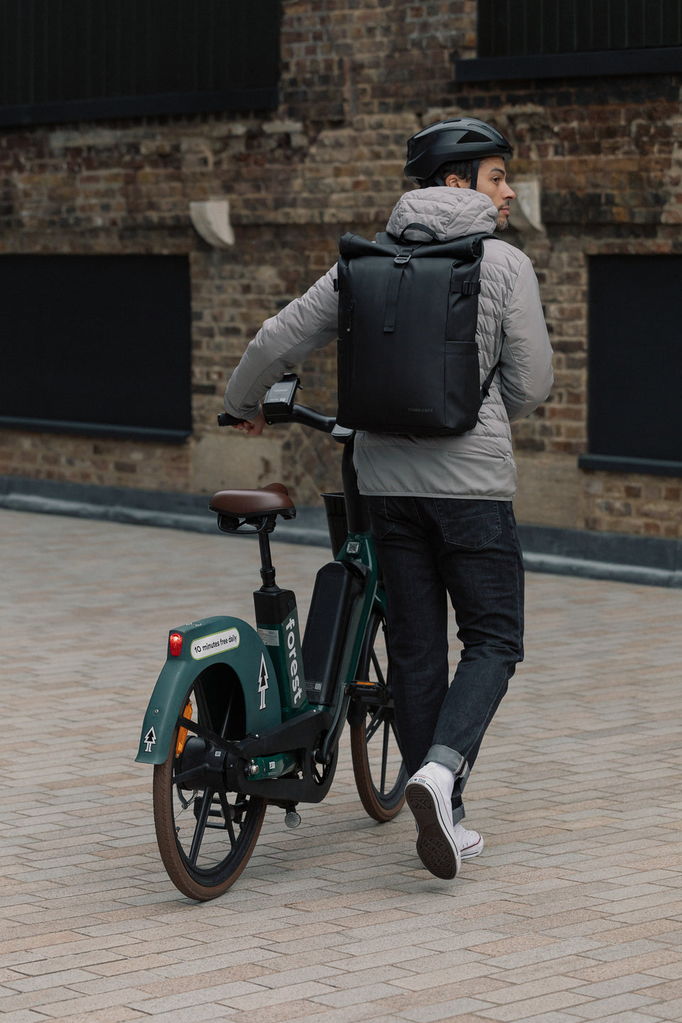 A man pushing an e-bike down the street whilst wearing a black backpack on his back