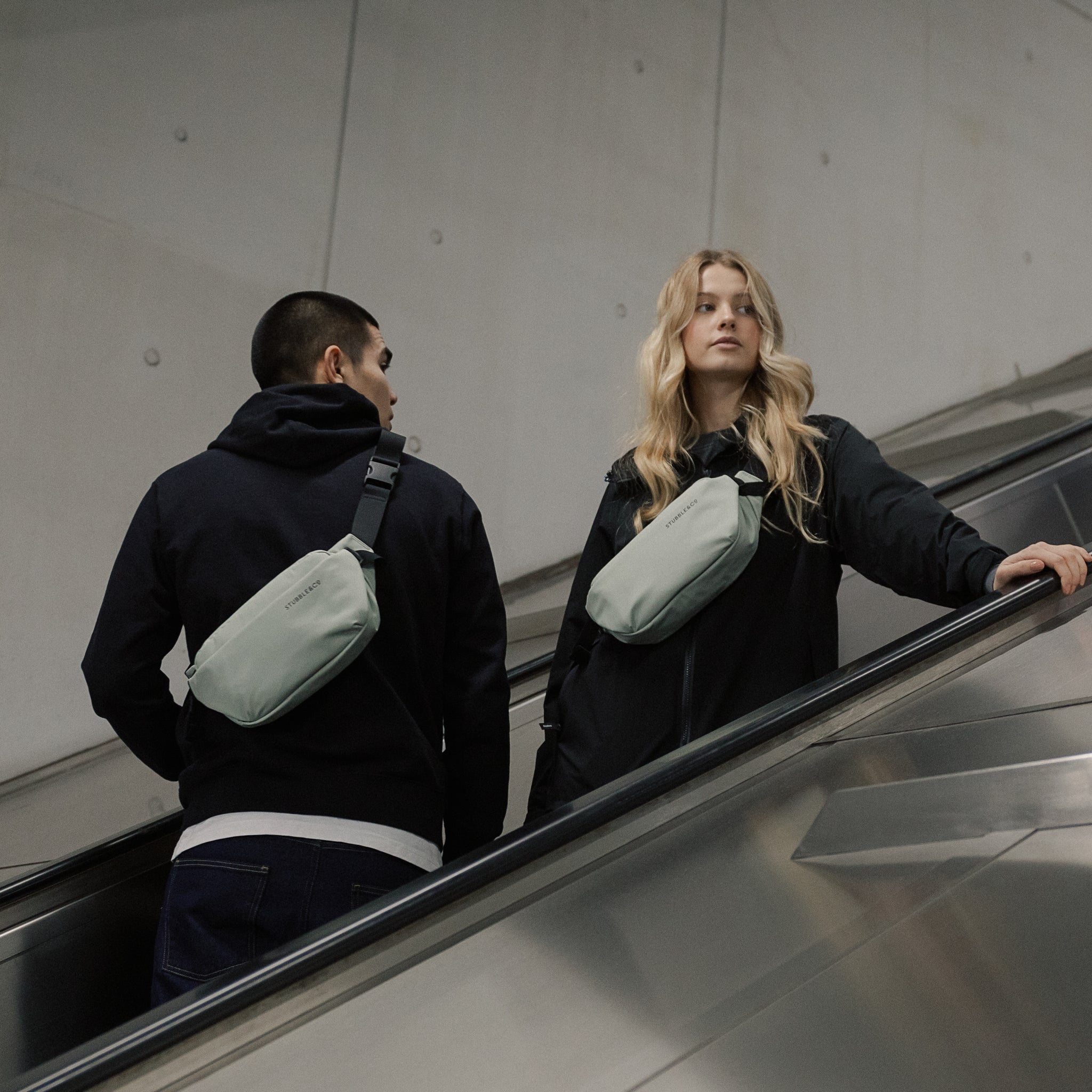 A man and a woman with a Matcha green crossbody bag on as they go down as escaltor