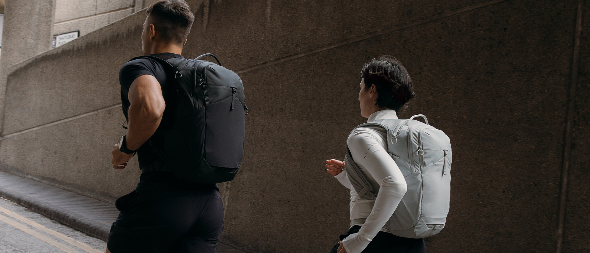 A man and woman running along a road and both wearing ash and charcoal motion backpack 20l on their backs