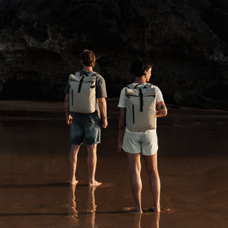 Man and woman stood on beach, man wearing The Roll Top in Sand, the woman wearing The Roll Top Mini in Sand