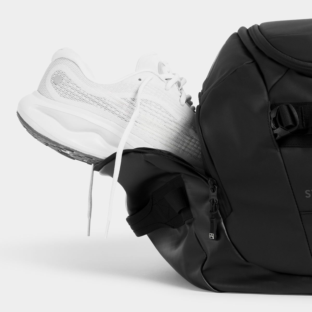 A white athletic sneaker is shown inserted into or pulled from a black backpack, photographed on a clean light gray studio background.