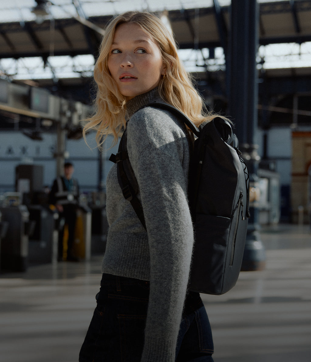 A woman walking up to the train station ticket barriers wearing a midnight roll top 15l on her back