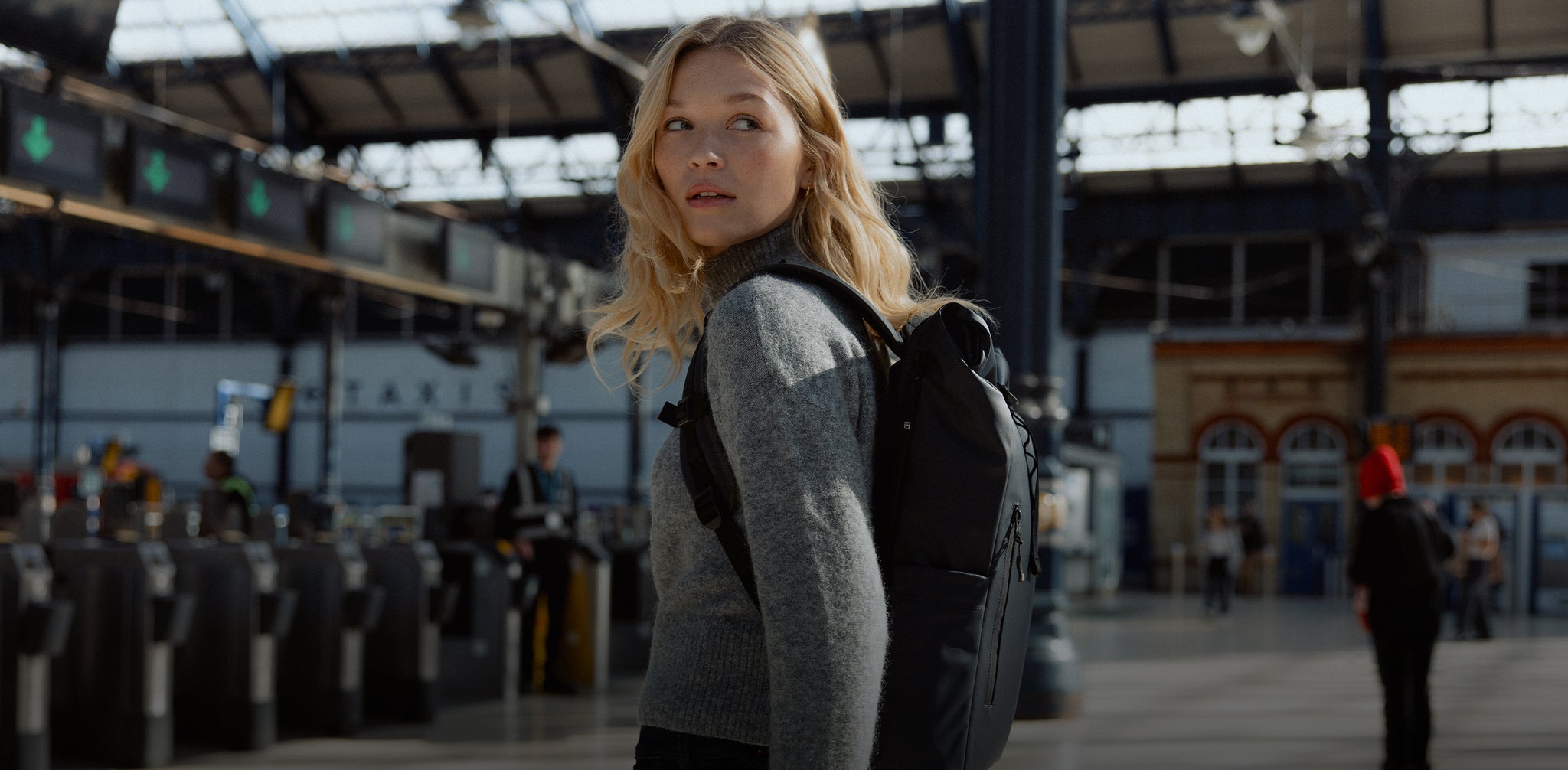 A woman walking up to the train ticket barrier with a midnight Roll Top 15L on her back- desktop
