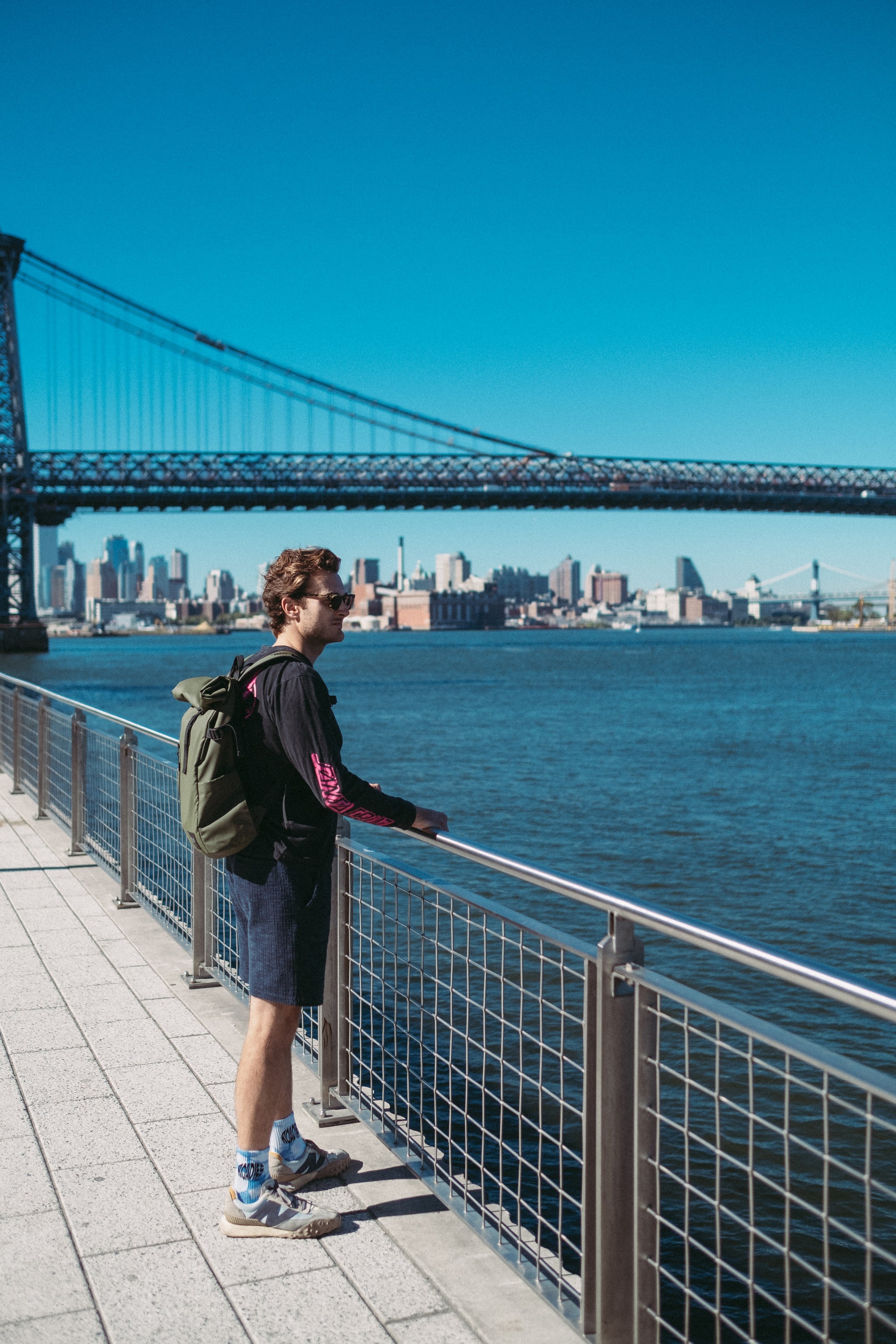 A man standing in view with new york city in the background wearing a green rucksack on his back