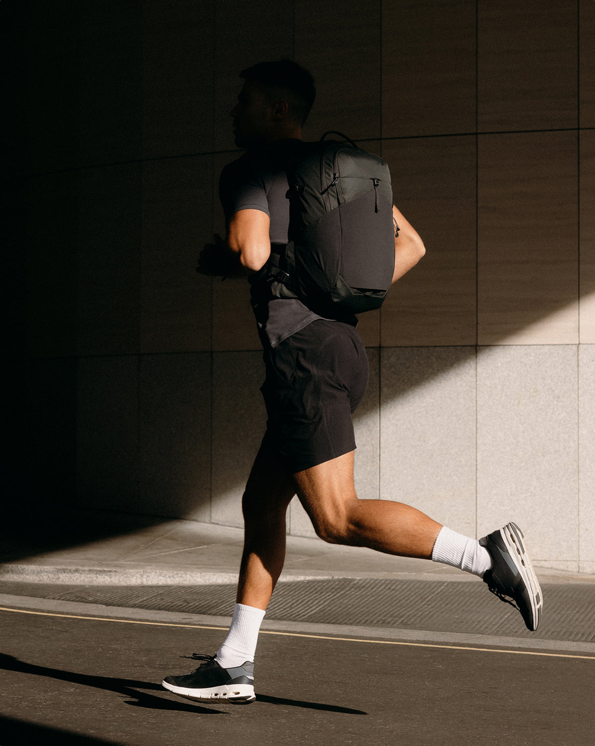 A man running outdoors on a sunny street, shown from behind wearing a black athletic backpack, black shorts, a dark gray t-shirt, white socks, and black running shoes against a tiled wall.