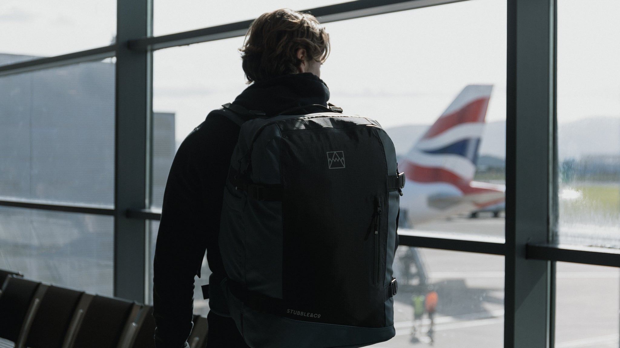A man walking through the airport with airplanes in the background with a tasmin blue adventure bag on his back