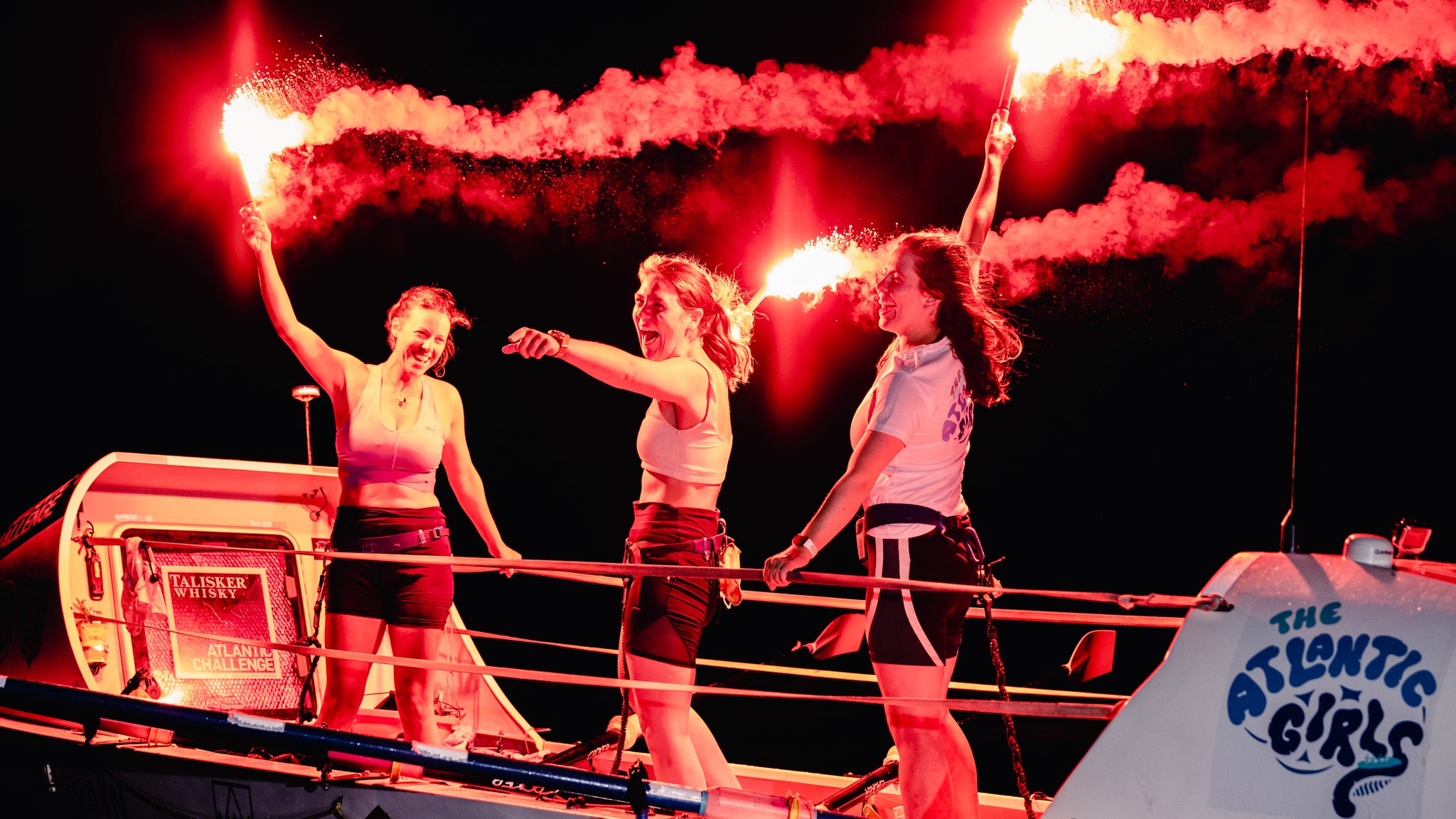 three woman holding red flares and cheering on a rowing boat