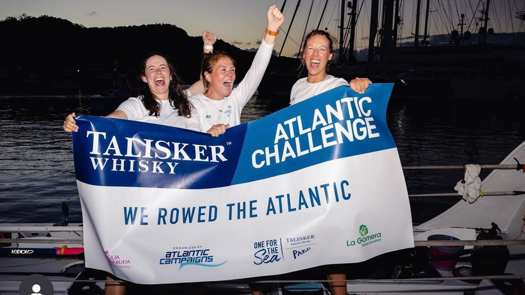 Three woman cheering with a sign showing they have completed a row across the Atlantic