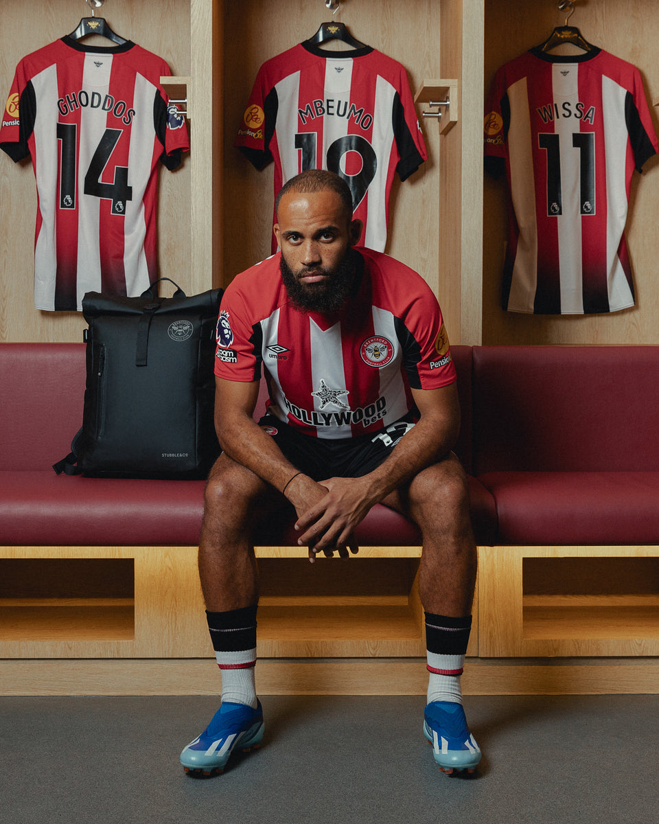 football player sitting on the changing room bench looking at camera with a black roll top next to him