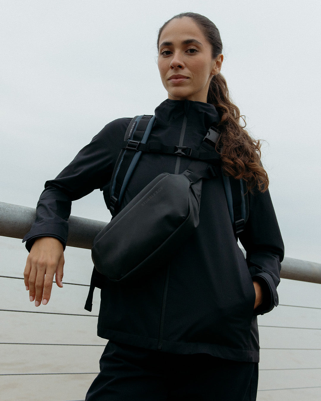 A woman leaning up against a fence with a crossbody on her front and a blue roll top backpack on her back with the chest straps done up