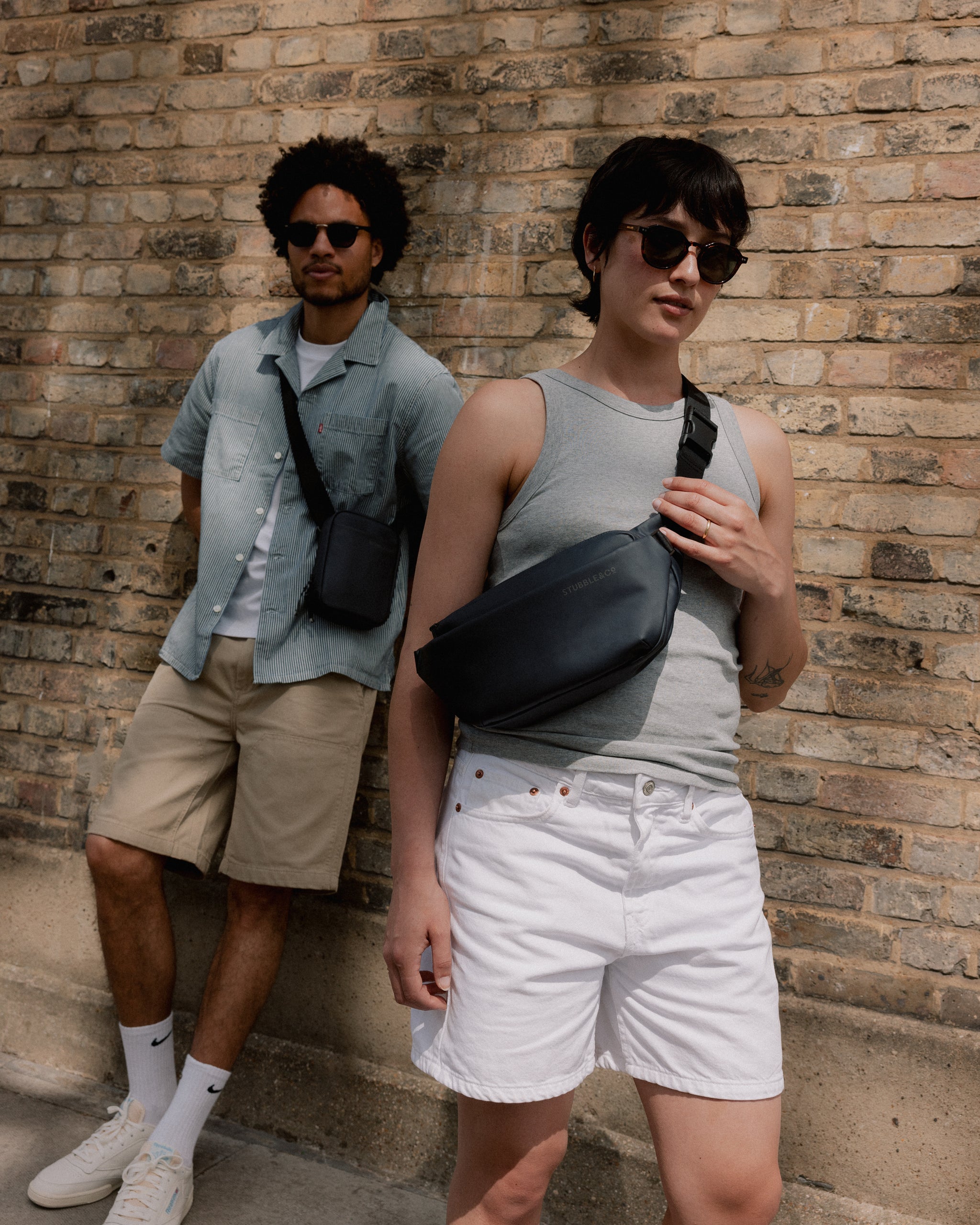 A woman wearing a midnight crossbody on her front with a man leaning against the wall wearing a midnight blue shoulder bag