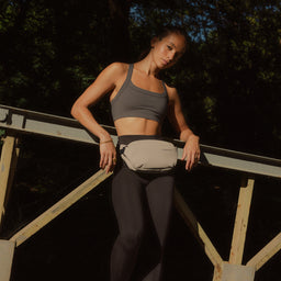 A woman leaning against a fence wearing a Sand crossbody across her waist
