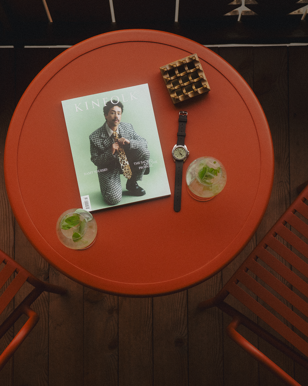 Round red table with a magazine, watch, and drinks on a dark tiled floor.