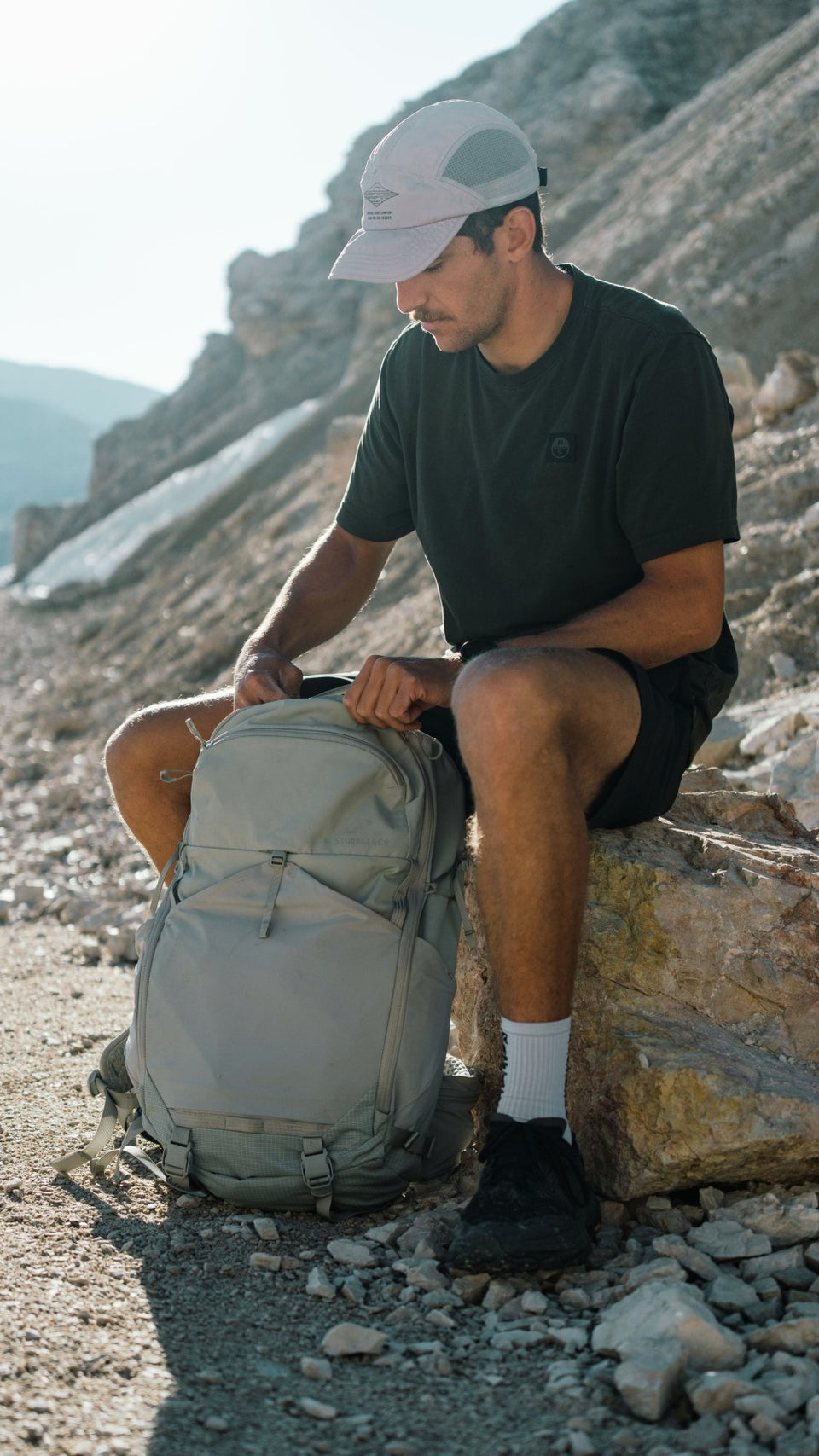 A man sitting on a rock in a rocky outdoor mountain setting, wearing a black t-shirt, black shorts, white socks, and a light gray cap while adjusting a large light gray hiking backpack.
