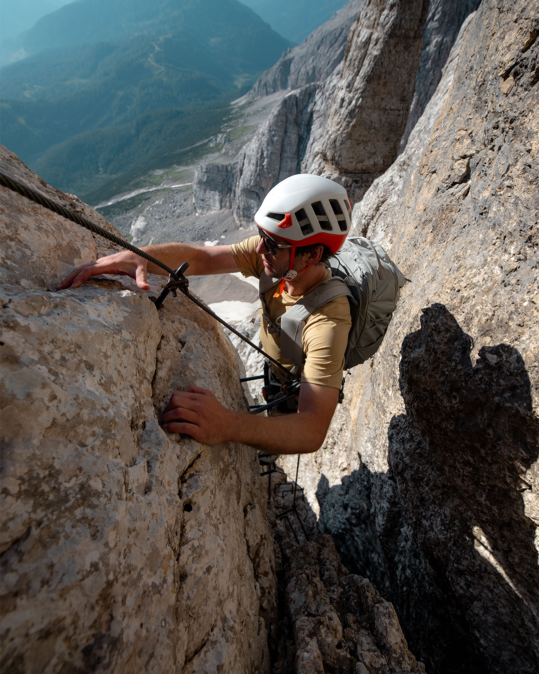 A rock climber wearing a white helmet and safety harness scales a steep, sunlit limestone cliffside with a deep mountain valley in the background.