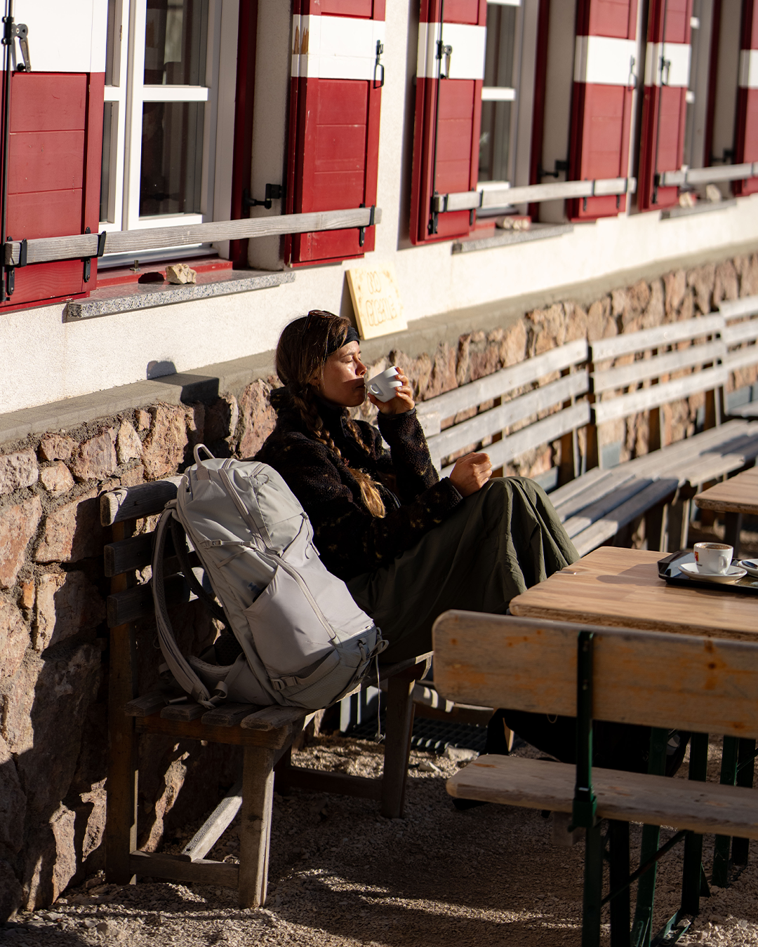 A woman sits on a wooden bench at an outdoor cafe, drinking from a white cup. A light gray backpack rests on the bench beside her.