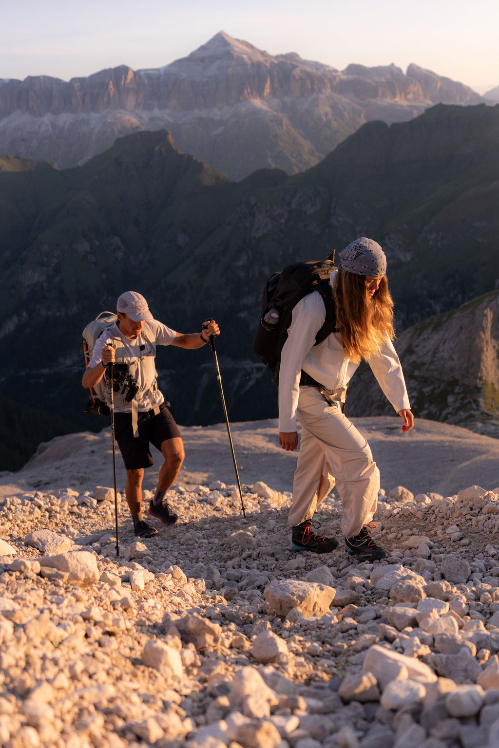 Two hikers with backpacks and trekking poles ascend a rocky mountain ridge, with rugged peaks in the background during golden hour.