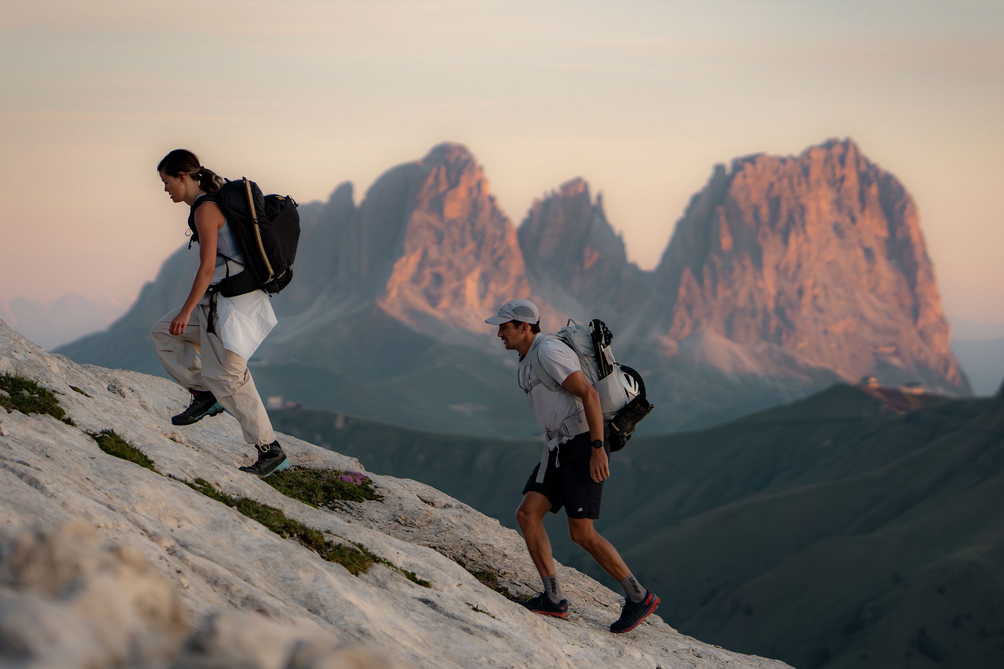 Two hikers with backpacks traverse a rocky mountain slope with dramatic peaks in the background during golden hour.