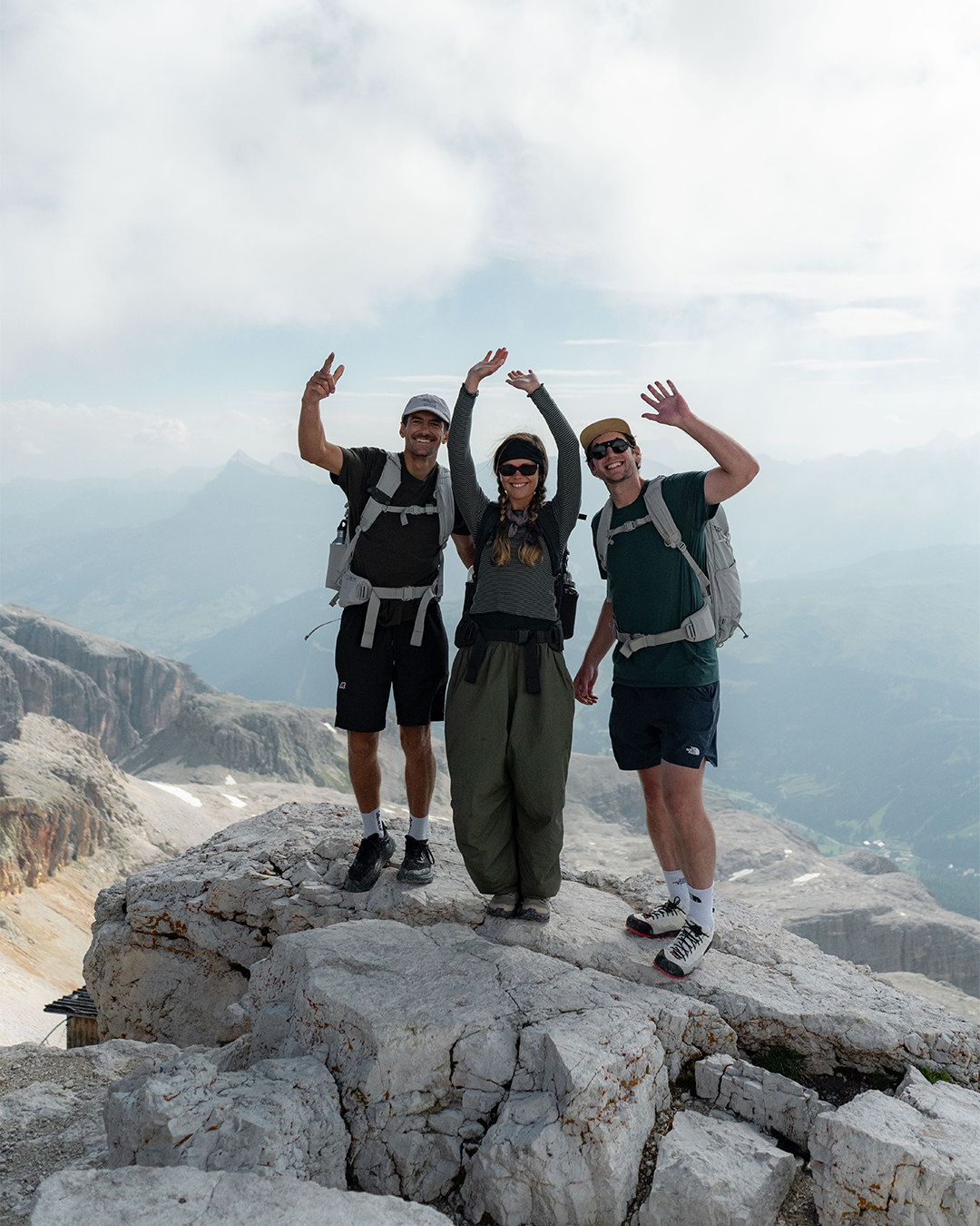Three hikers posing together on a rocky mountain summit with a scenic alpine landscape in the background under a cloudy sky.