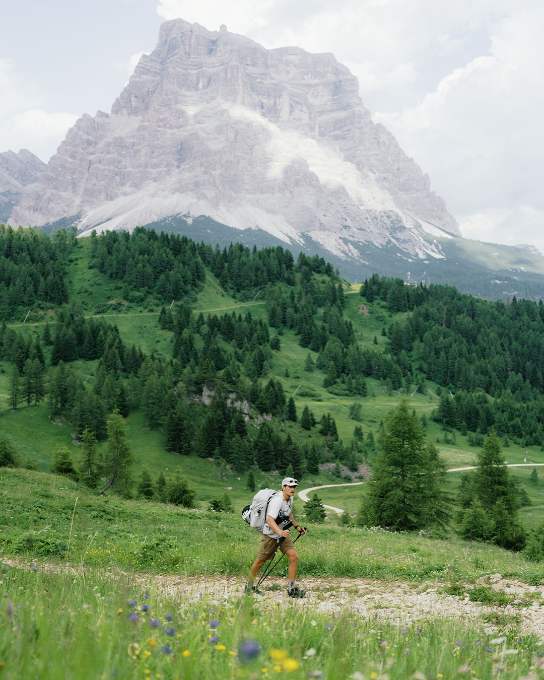 A hiker with a backpack walks along a dirt path in a grassy mountain meadow, with a large rocky peak and green forest in the background.
