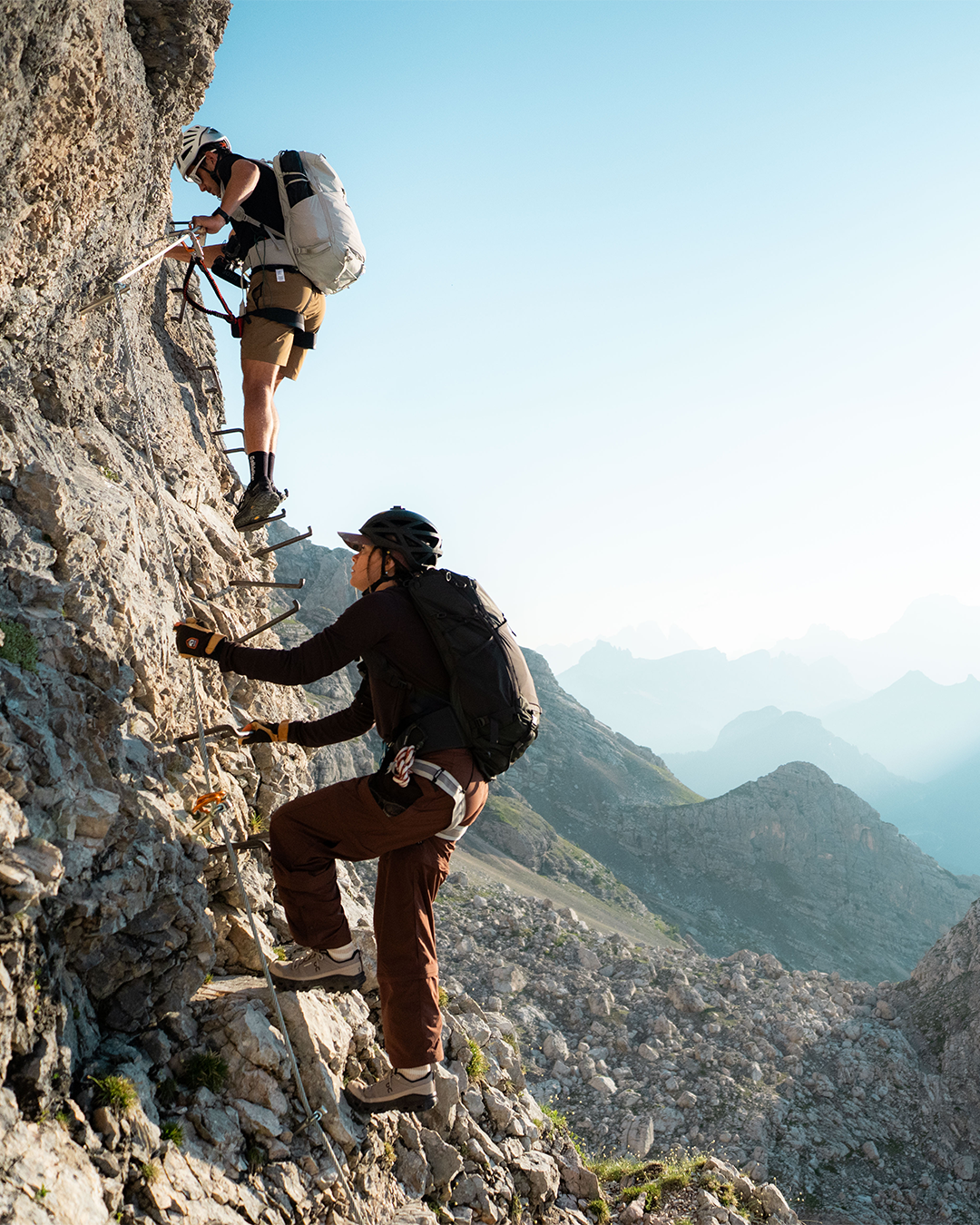 Two mountain climbers with backpacks and helmets ascend a steep rocky cliff using metal ladder rungs, with a vast mountain range visible in the background under a clear blue sky.