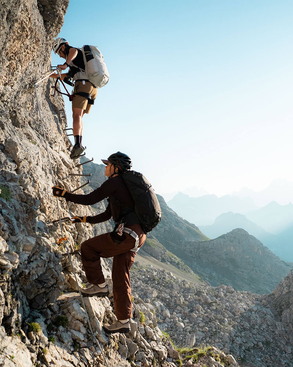 Two mountain climbers with backpacks and helmets ascend a steep rocky cliff using metal ladder rungs, with a vast mountain range visible in the background under a clear blue sky.