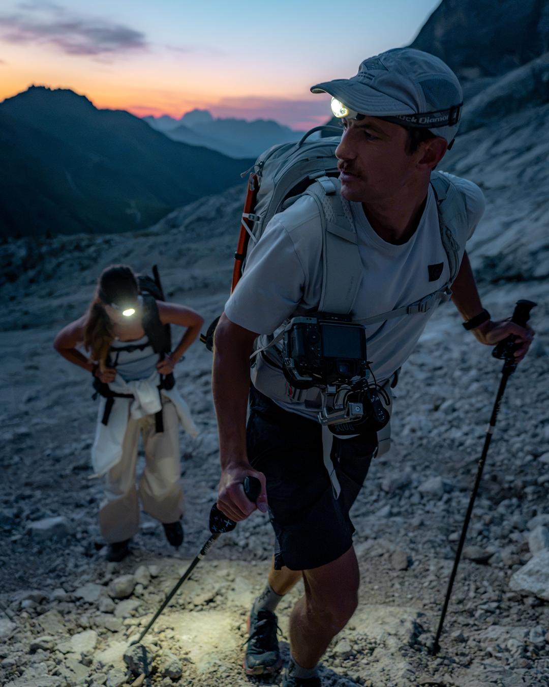 A man hiking in a rocky mountain landscape at dusk, wearing a light gray cap with a headlamp, a light gray t-shirt, dark shorts, and a backpack with a mounted camera. Another hiker is visible in the background.