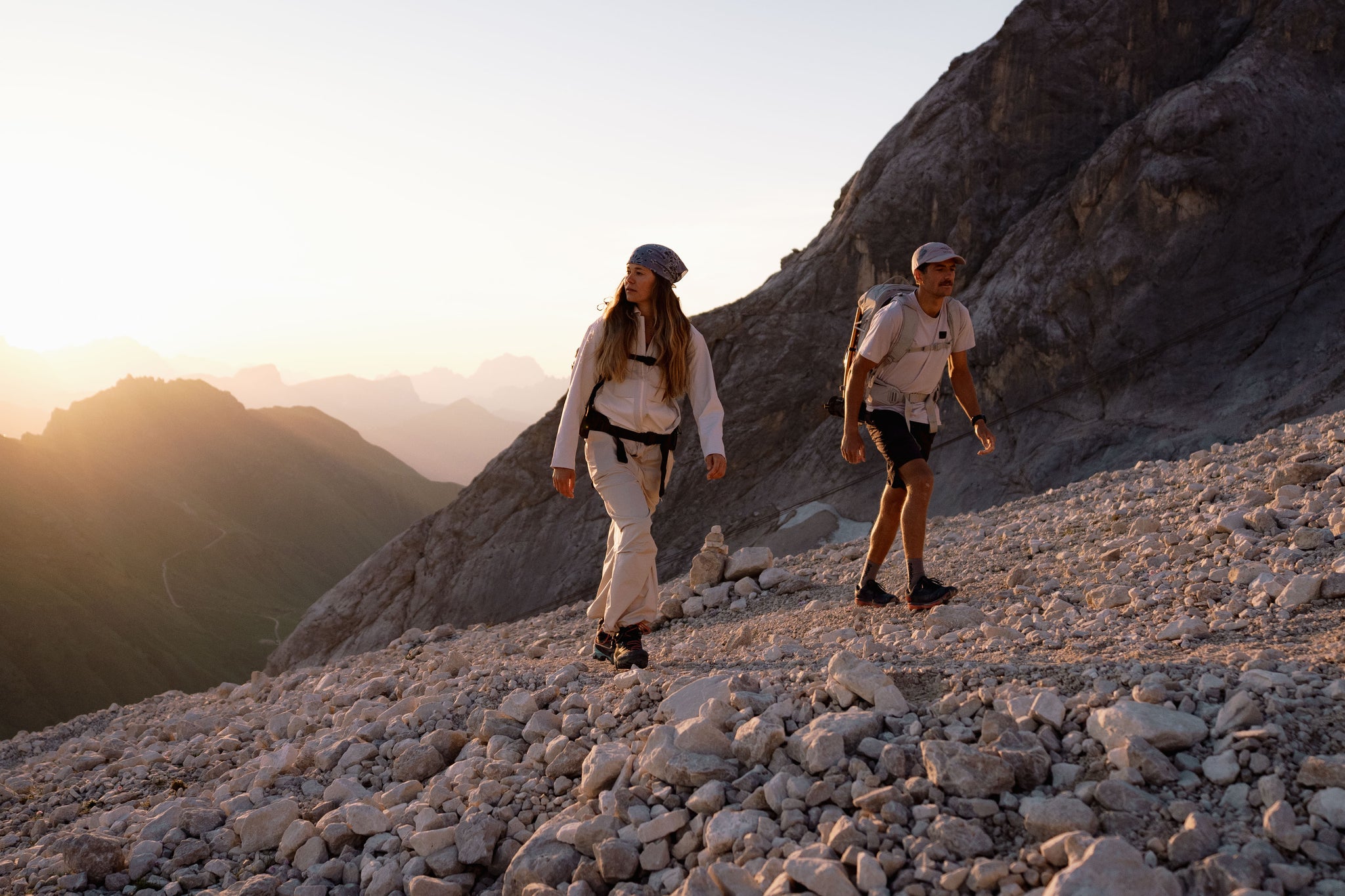 Two hikers walking on a rocky mountain slope during golden hour, with a large rock face and distant mountain ridges in the background.