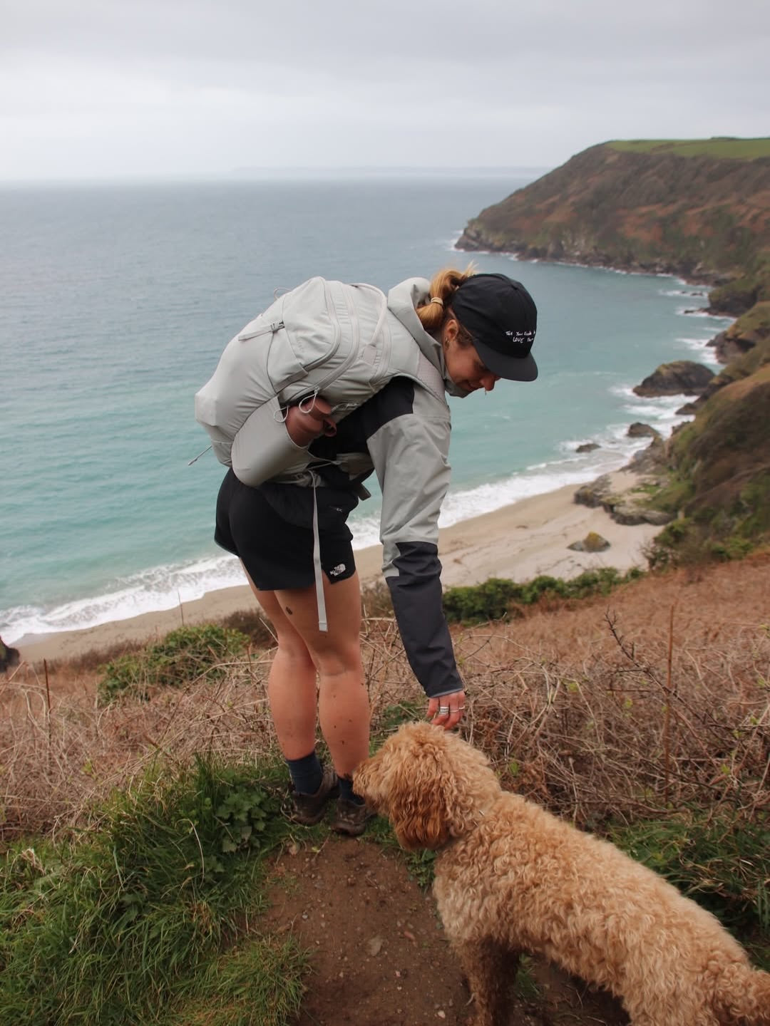 Woman in a cap and grey jacket crouching to stroke a curly golden dog on a clifftop path above a sandy beach