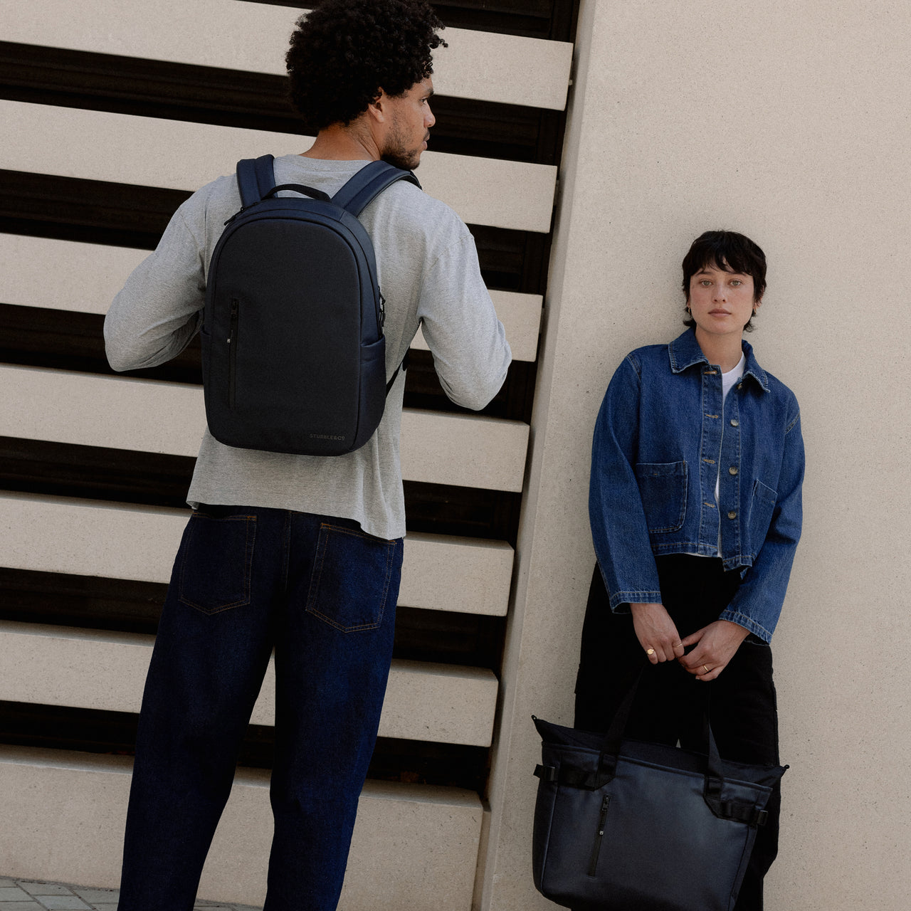  A man wearing an Everyday Backpack 15L in Midnight Blue on his back standing next to a woman holding a Laptop Tote Bag