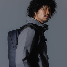 A studio shot of an Everyday Backpack 15L in Midnight Blue showing a man standing side on wearing the bag on his back