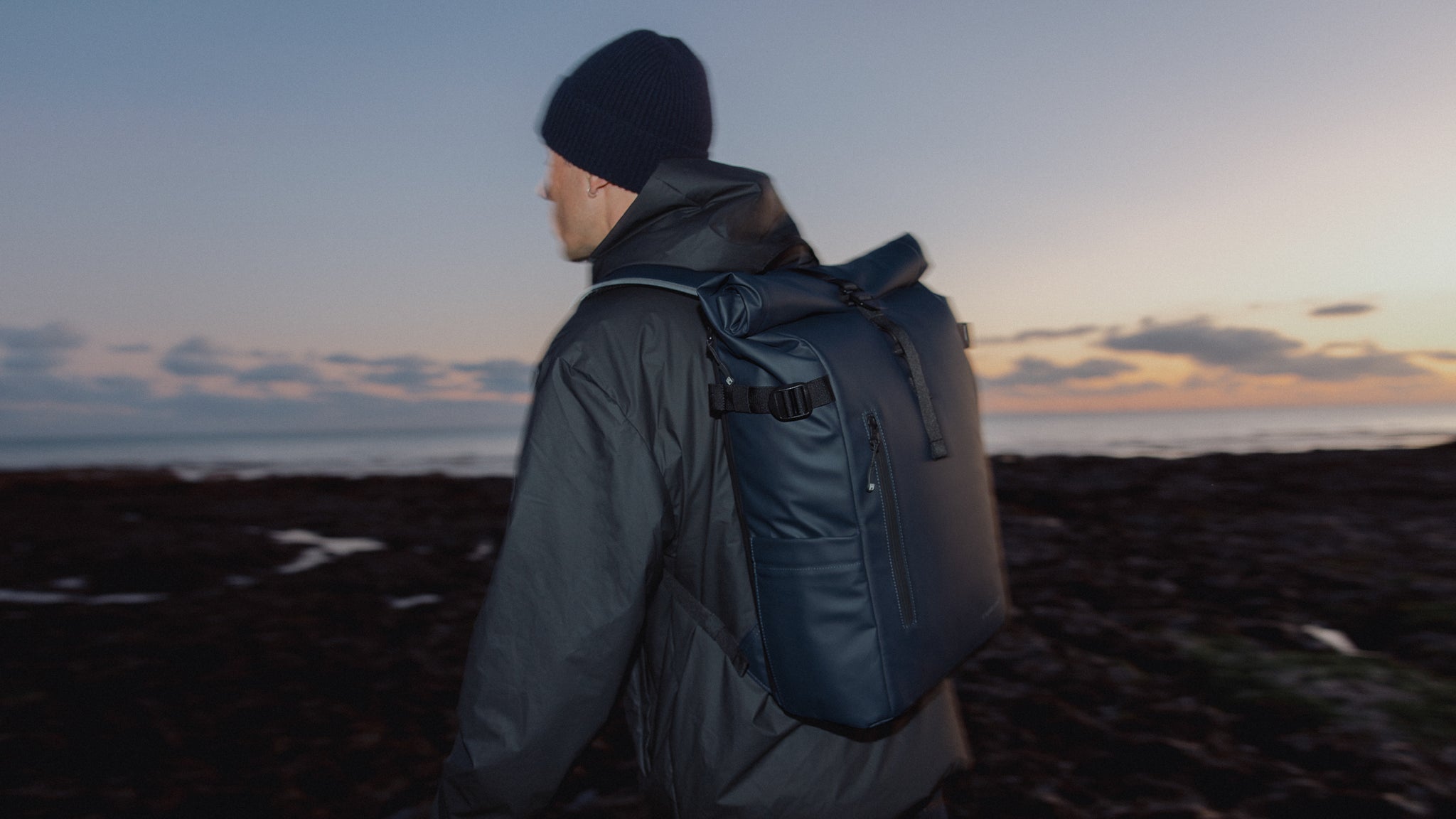 A man standing on the beach at sunset wearing a Roll Top 20L in Midnight Blue on his back
