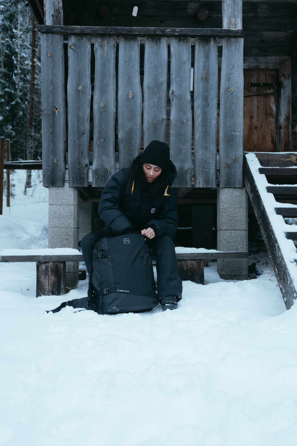 A woman sitting in front of a cabin in the snow packing her black backpack