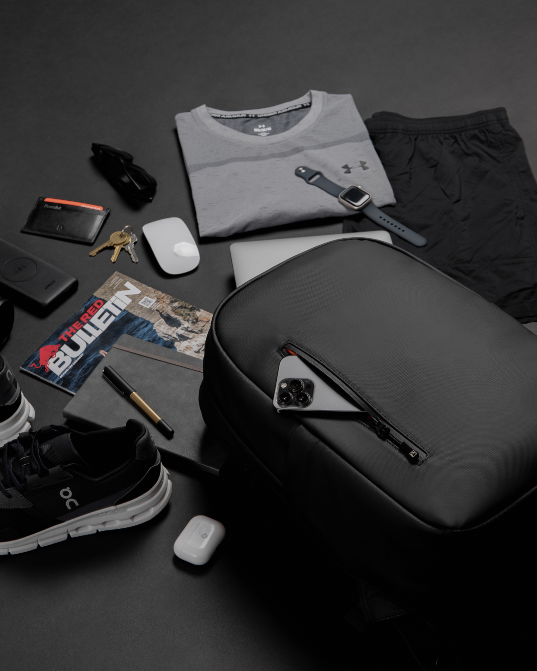 Flat lay of the Everyday Backpack in All Black with essentials including a t-shirt, trainers, wallet and tech accessories. 