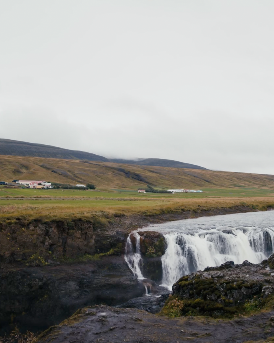 A grassy landscape with foggy skies, rolling hills and a small waterfall and river