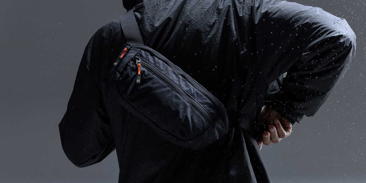 A person wearing a black waterproof jacket and black pants, shown from the back while wearing a black crossbody sling bag with orange zipper pulls. The image is set in a dark, moody environment with a rain-like effect.