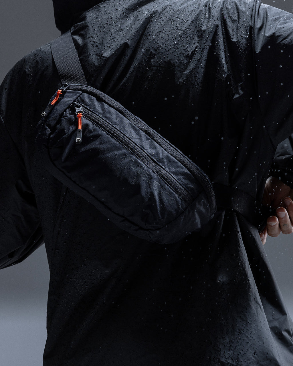 A black waterproof crossbody bag worn across the chest of a person in a black rain jacket, shown from behind in a dark, moody setting with water droplets.