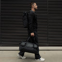 A man walking down the street wearing a roll top backpack on his back and holding a gym duffel 25l in All Black at his side.