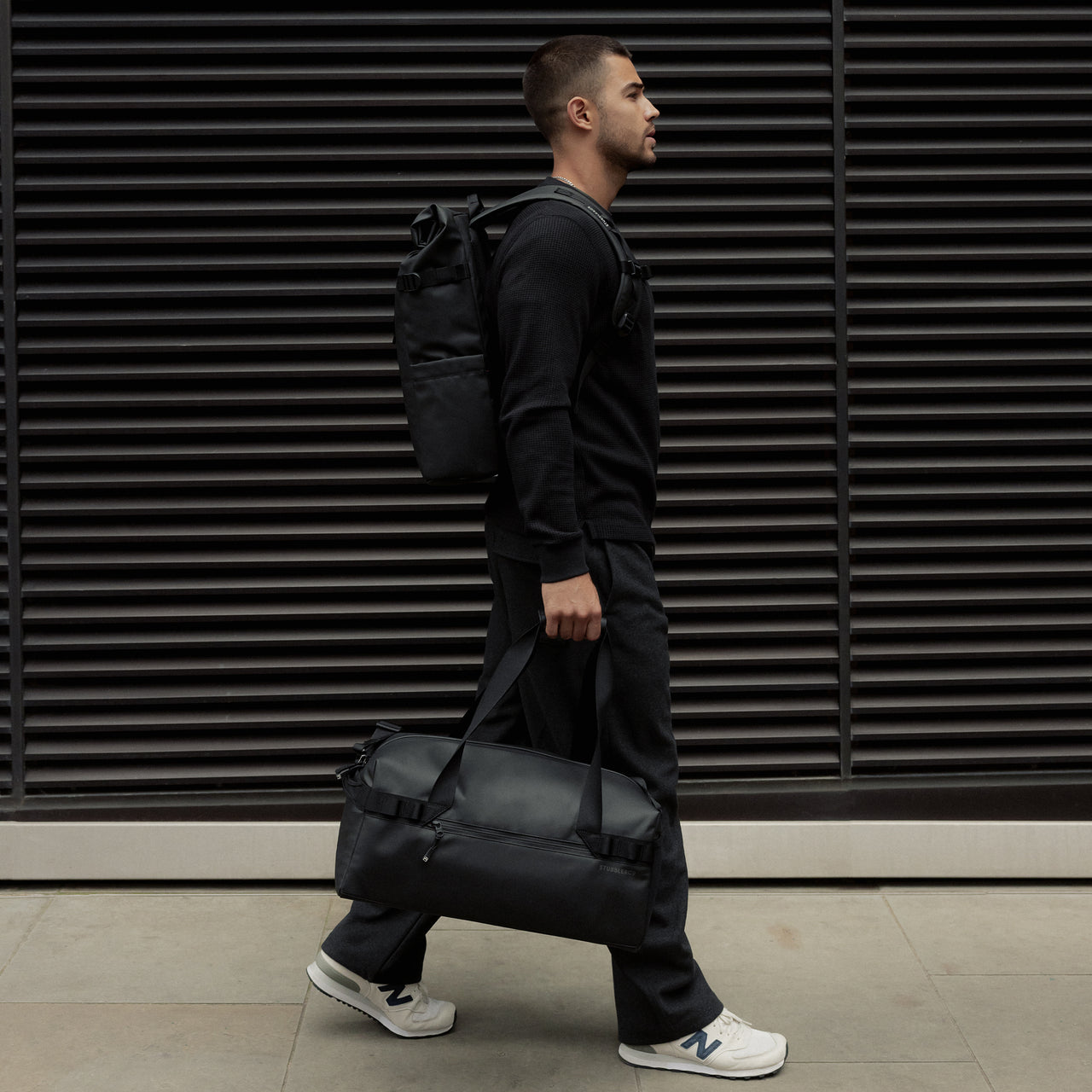 A man walking down the street wearing a roll top backpack on his back and holding a gym duffel 25l in All Black at his side.