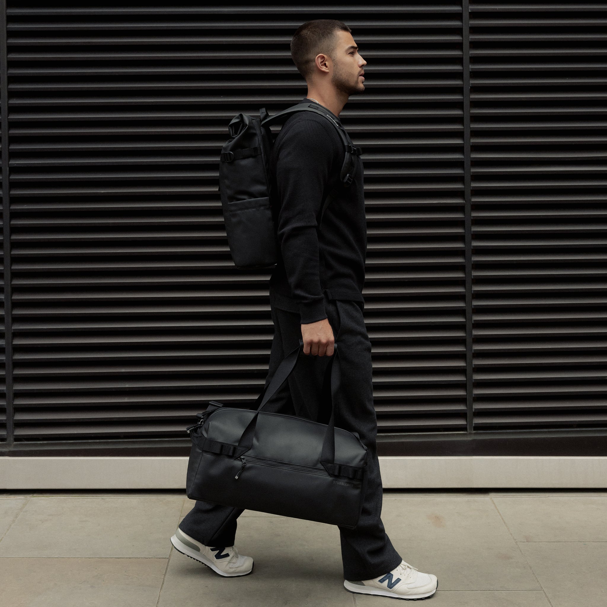 A man walking down the street wearing a roll top backpack on his back and holding a gym duffel 25l in All Black at his side.