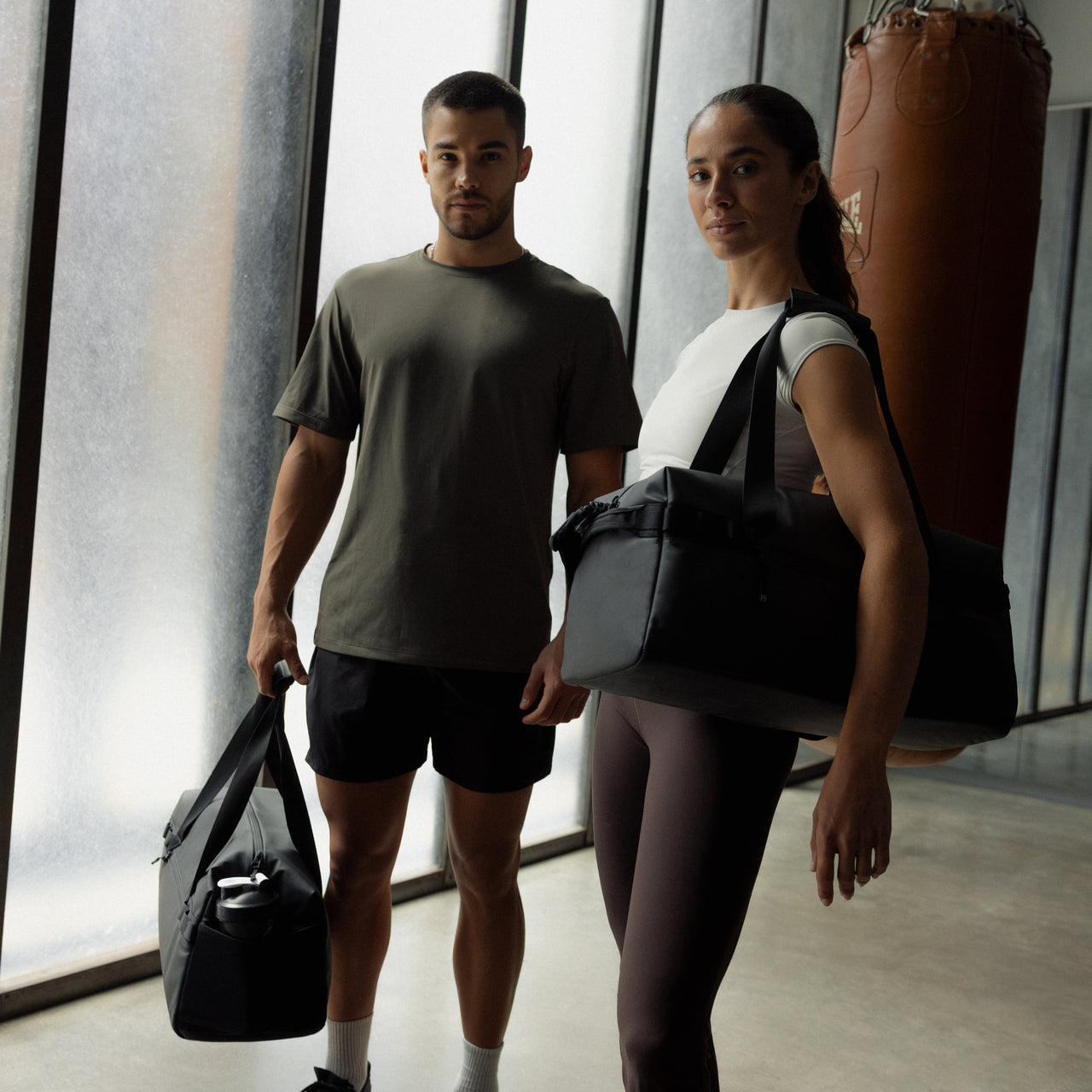 A man and woman standing in a gym with a boxing bag holding a gym duffel 25L in All Black